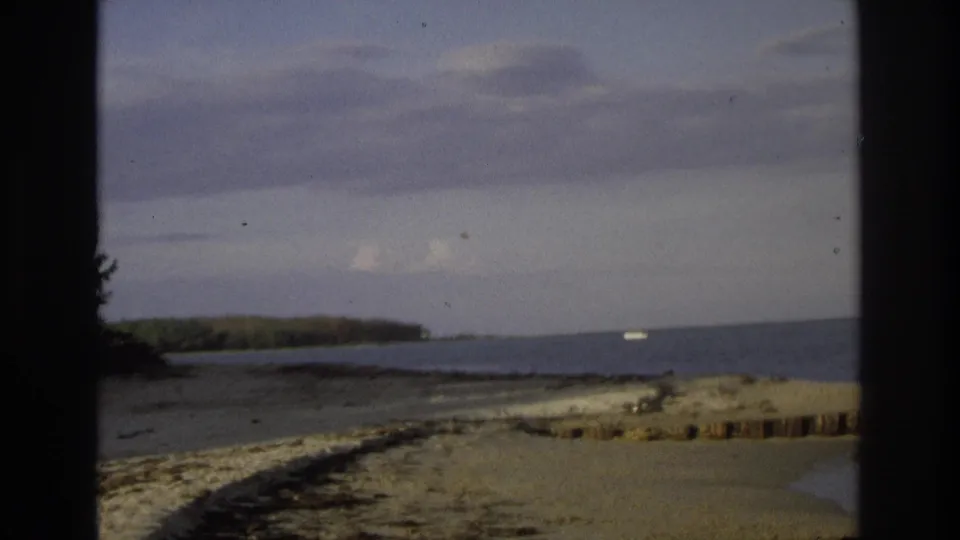 A 1968 home-movie view of Ocean City beach and ocean, showing sand in the foreground, a distant wooded shore, cloudy sky, and a small boat offshore.