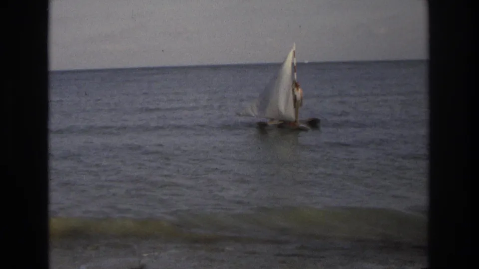 Small white-sailed boat on the ocean near Ocean City, New Jersey in 1968, with a distant person aboard and low waves under a muted sky.