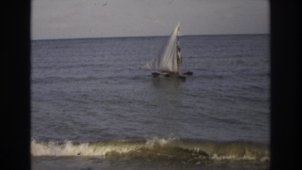 Small sailboat with a white triangular sail on calm ocean water near breaking surf.