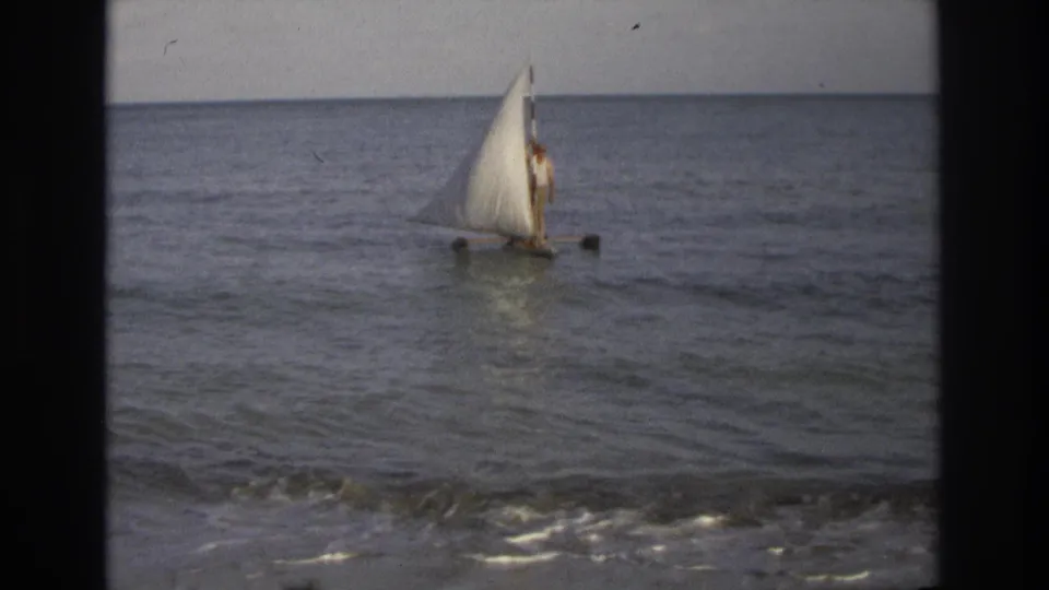 1968 8mm coastal frame from Ocean City showing a person on a small white-sailed boat beyond gentle beach surf.