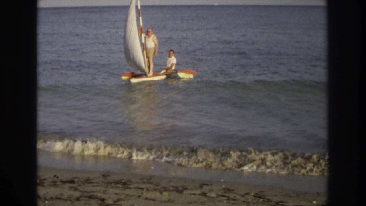 Two men on a small sailboat near shore with a white sail and low waves.