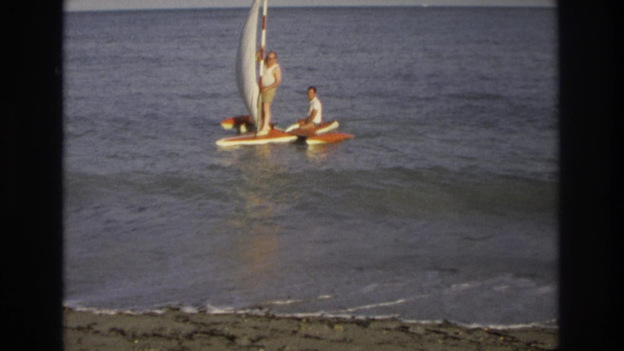 Small sailboat with two men floating on calm water near the beach.