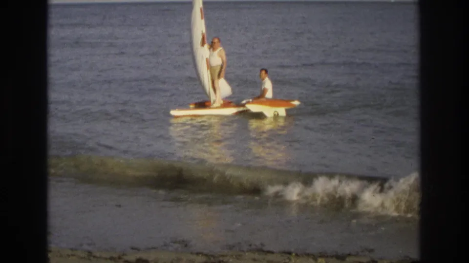 A home-movie frame from Ocean City shows one man standing by the sail and another seated while a small wave breaks nearby.