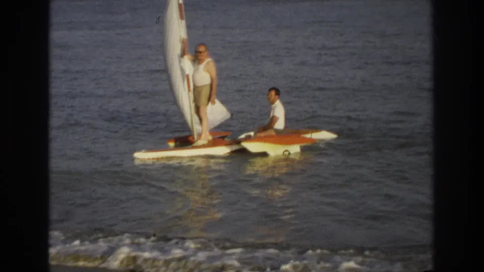 Closer 1968 Ocean City frame showing one man holding the sail while another sits on the small orange-and-white boat near shore.