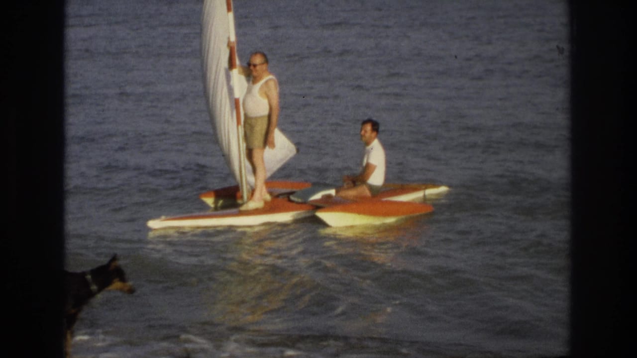 Two men on a small sailboat in ocean water as a black dog enters the shoreline foreground.