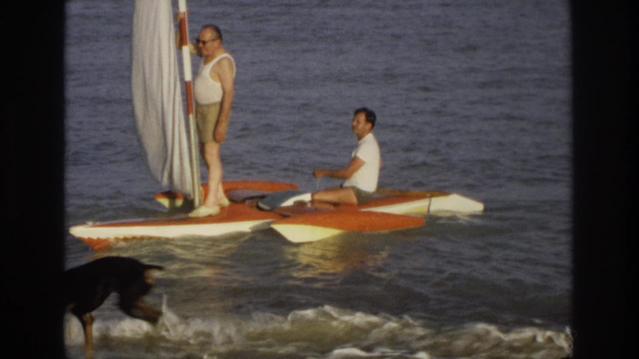 1968 Ocean City frame of two men on a small catamaran-style sailboat as a black dog passes through the foreground surf.