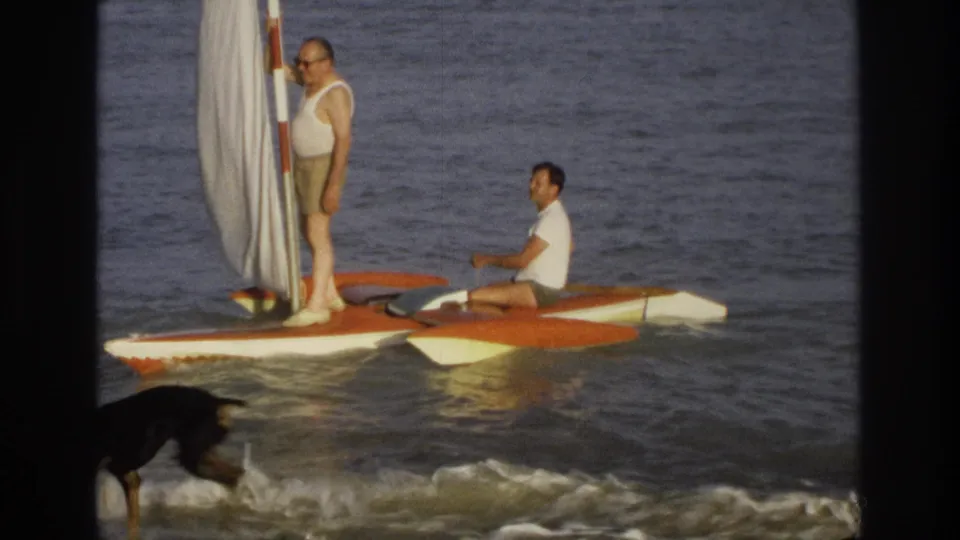1968 Ocean City frame of two men on a small catamaran-style sailboat as a black dog passes through the foreground surf.