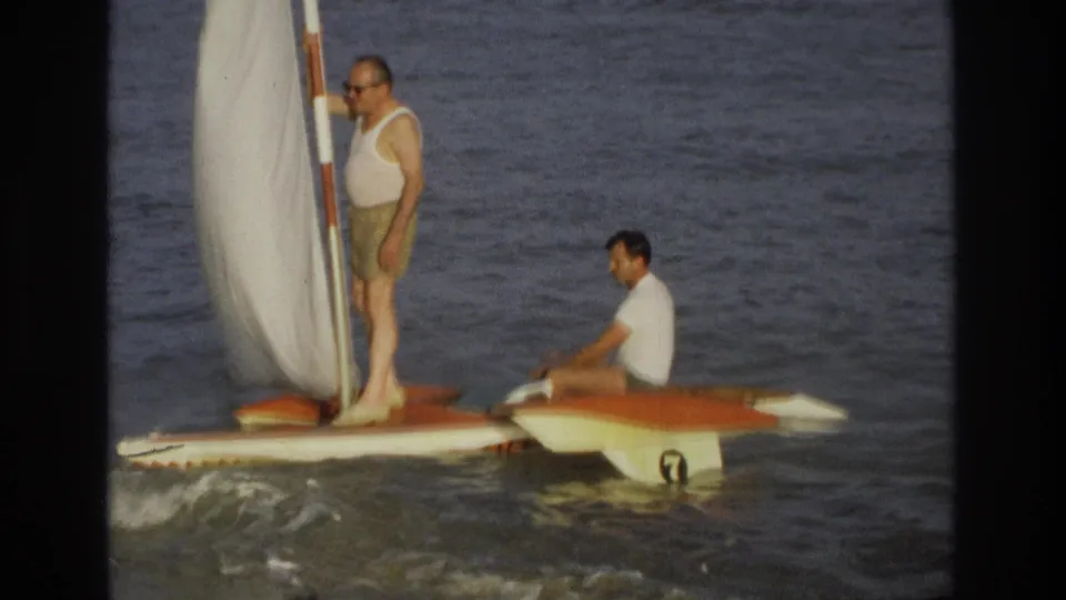 1968 Ocean City home-movie frame showing a closer side view of two men on a small sailboat, with surf and a hull number visible.