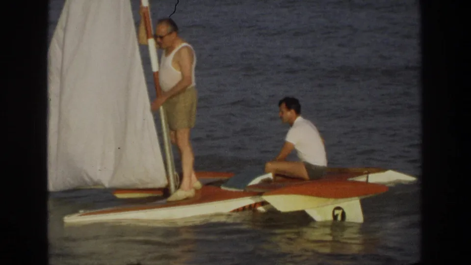 1968 Ocean City frame showing two men aboard a small orange-and-white sailboat with a white sail and visible hull marking.