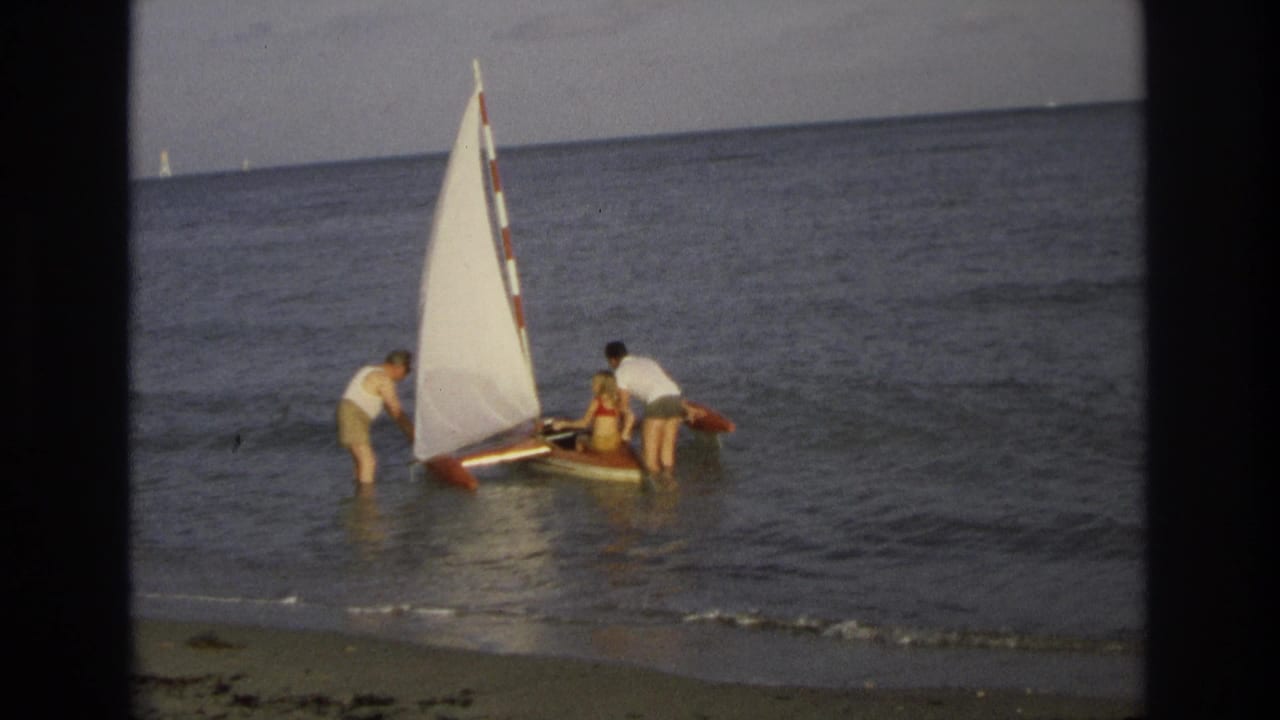 In Ocean City, New Jersey, 1968, three beachgoers help a small sailboat with a white sail in shallow surf.