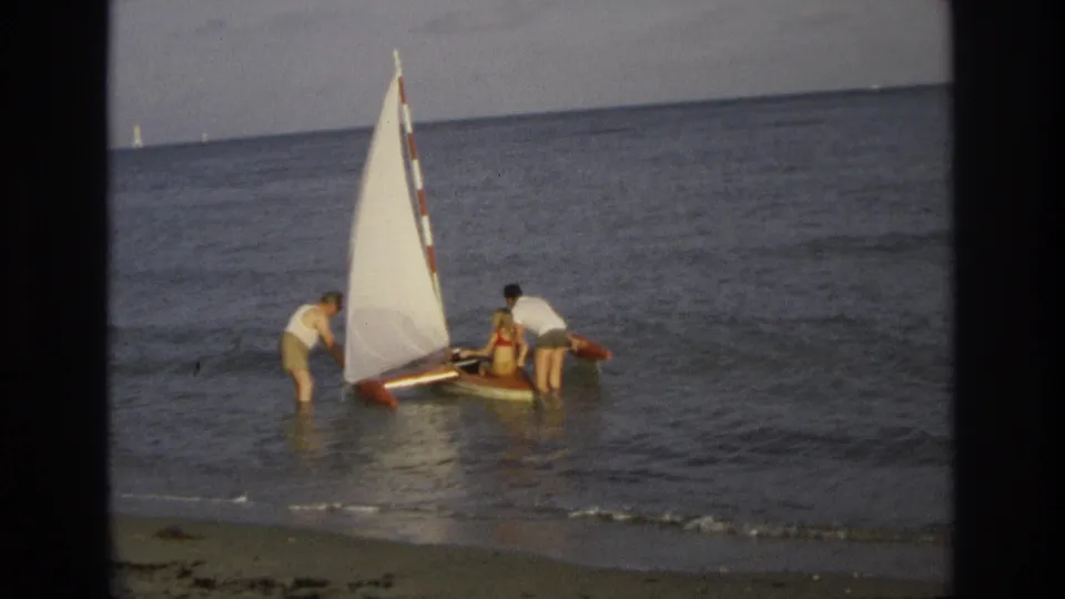In Ocean City, New Jersey, 1968, three beachgoers help a small sailboat with a white sail in shallow surf.