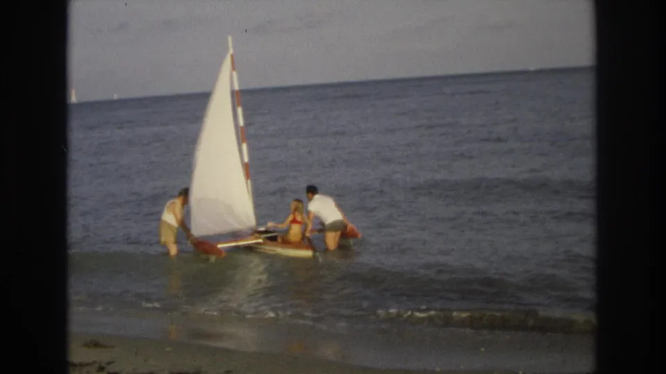 Three people in shallow Ocean City surf steady a small sailboat with a white sail and red trim in 1968 home-movie footage.