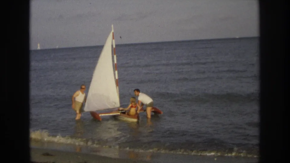 A home-movie frame shows beachgoers around a small sailboat in shallow Ocean City water, with the sail raised and waves nearby.