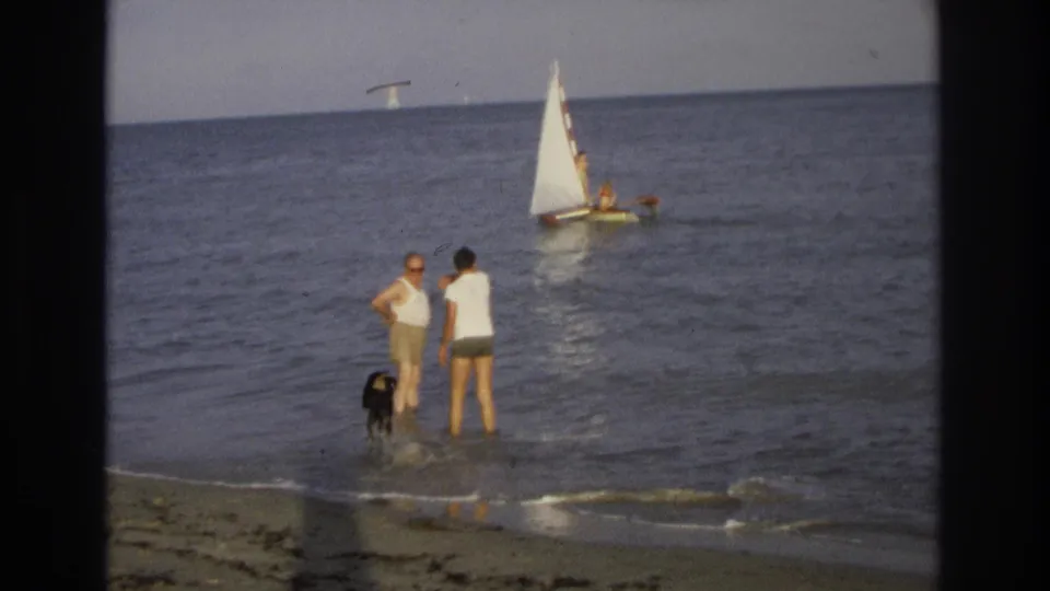 The couple stands in shallow ocean water near their black dog while a small white sailboat passes in the background at Ocean City, New Jersey.