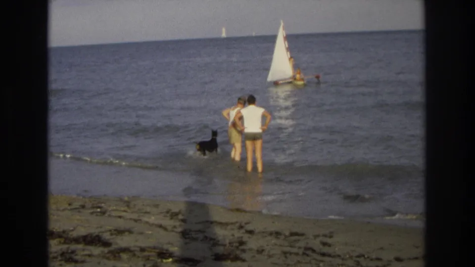 A 1968 home-movie frame shows two adults standing at the edge of the surf with a black dog nearby and a small sailboat on the water beyond.