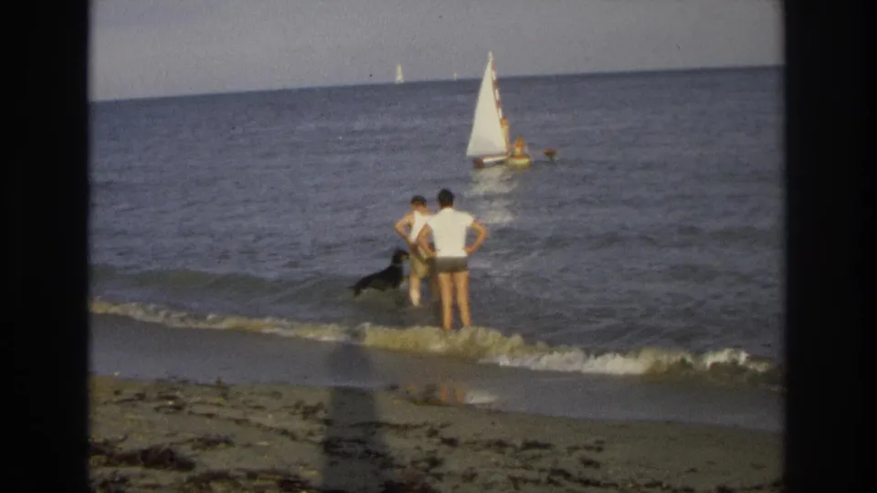 At the Ocean City shoreline, the adults stand together in shallow surf as their black dog moves near the breaking wave and a sailboat sits offshore.