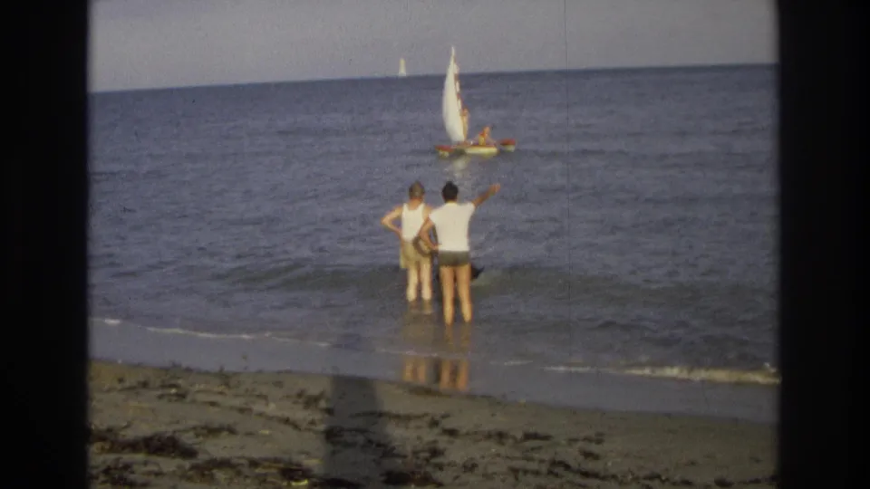 At the Ocean City shoreline, two men in white shirts stand in the surf as one points out toward a small sailboat on calm water.