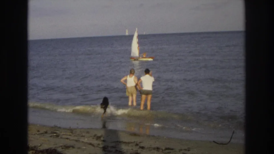 1968 Jersey Shore home-movie frame with a dog running through a small wave near two men standing at the edge of the ocean.