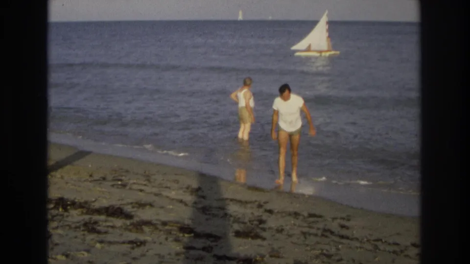 Ocean City beach frame showing one man walking toward the sand, another in the shallows, and a white-sailed boat crossing the background water.
