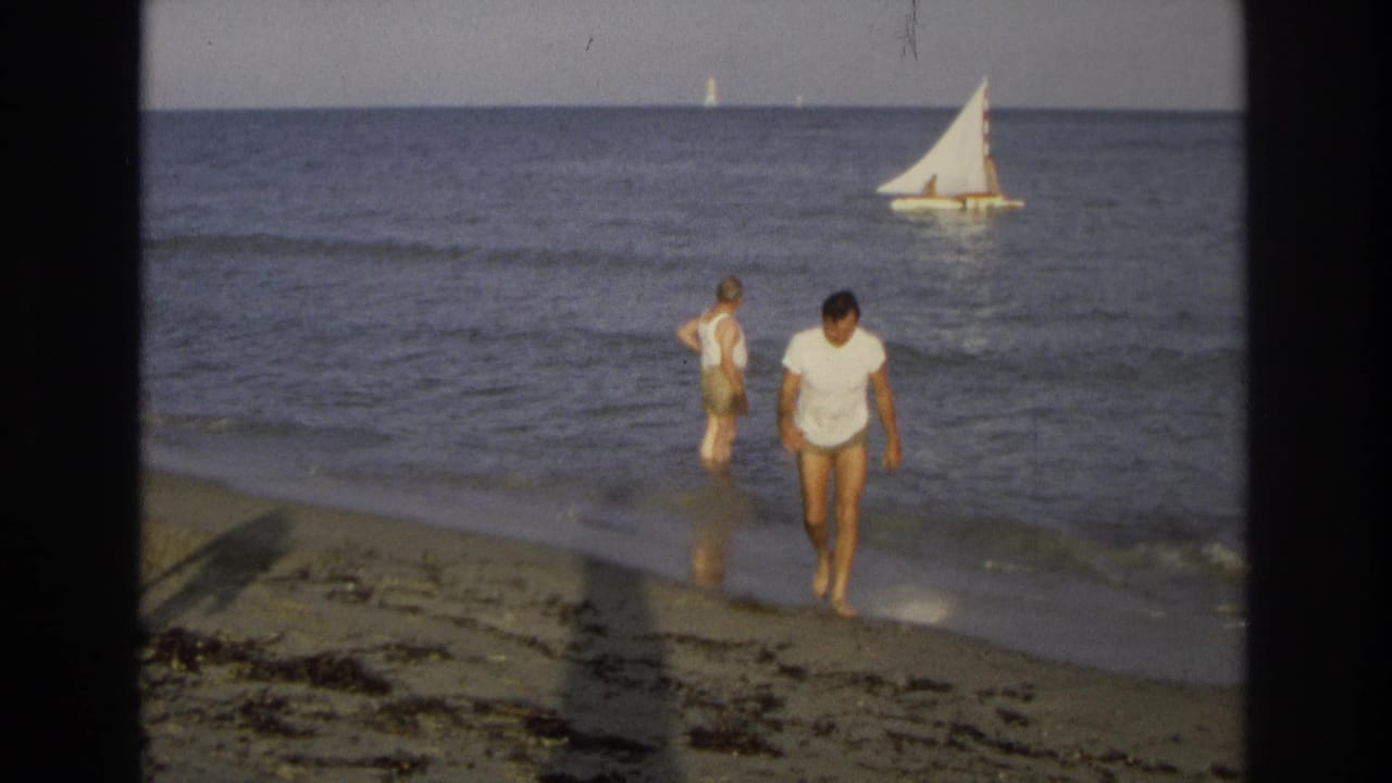 Two men walk near shallow surf as a small sailboat moves offshore.