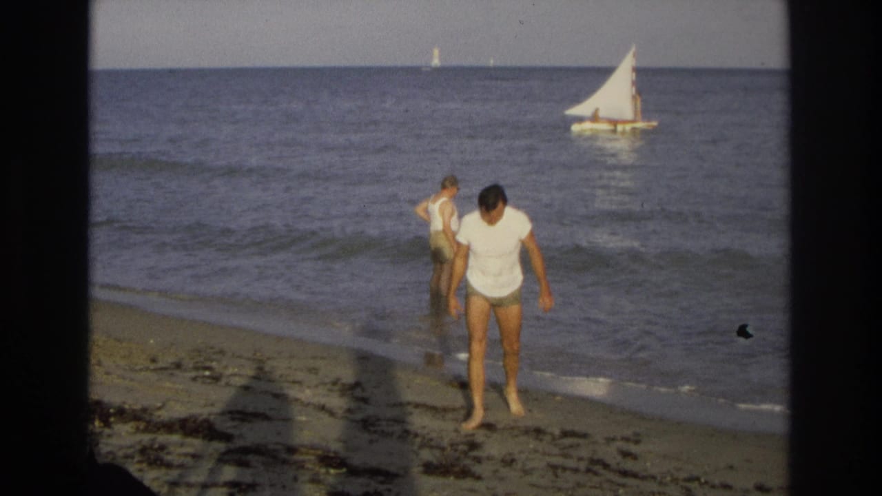 A man in a white shirt steps toward the camera beside waves and a distant sailboat.