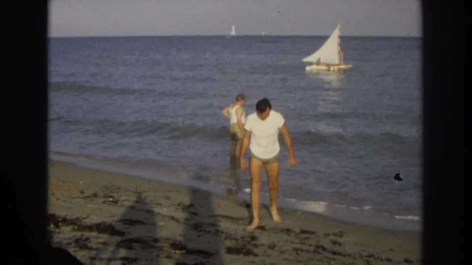 Man in a white shirt walks along the Ocean City shoreline with another beachgoer behind him and a sailboat offshore.