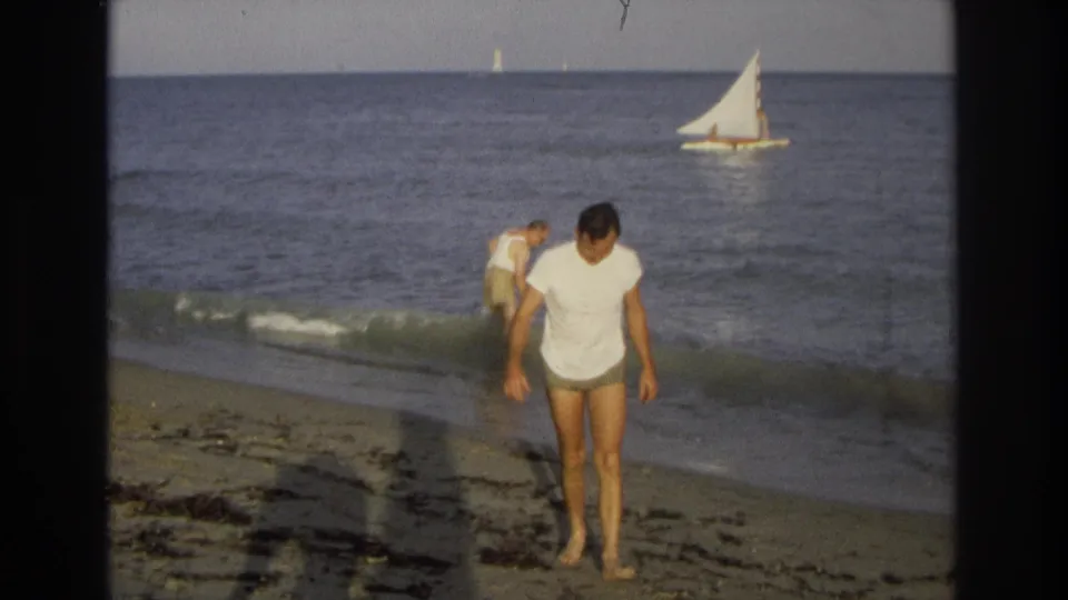 Ocean City beach frame showing a man walking barefoot in front as another bends at the waterline, with a sailboat in the distance.