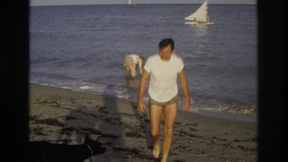 Close 8mm frame of a man walking past the camera on the Ocean City beach, with surf, seaweed, and a sailboat behind him.