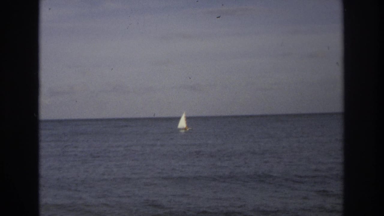 Distant sailboat with a white sail on the ocean under an overcast sky.