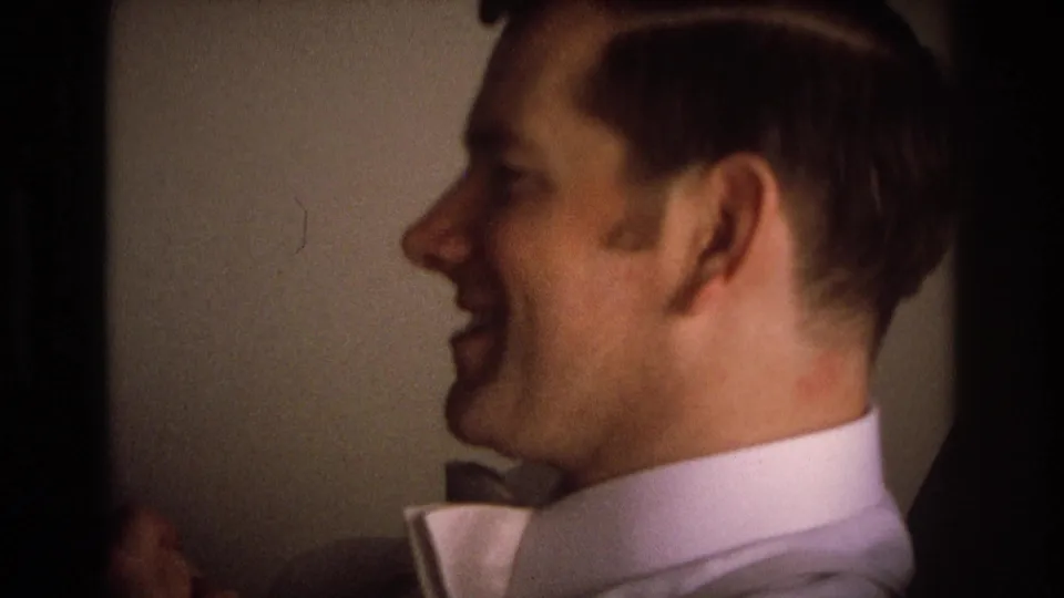 An adult man smiles in profile in 1968 home-movie footage, wearing a light collared shirt near a shaded interior or opening.