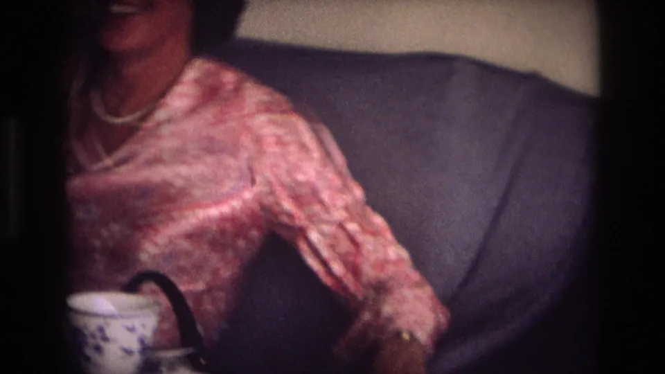In the 1968 Ocean City home movie, a woman in a pink patterned dress and pearl necklace laughs while holding a cup on upholstered seating.