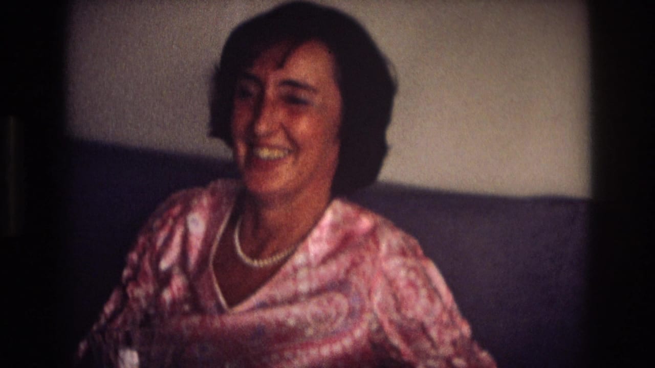 Woman in a pink patterned blouse smiles while seated on a dark couch.