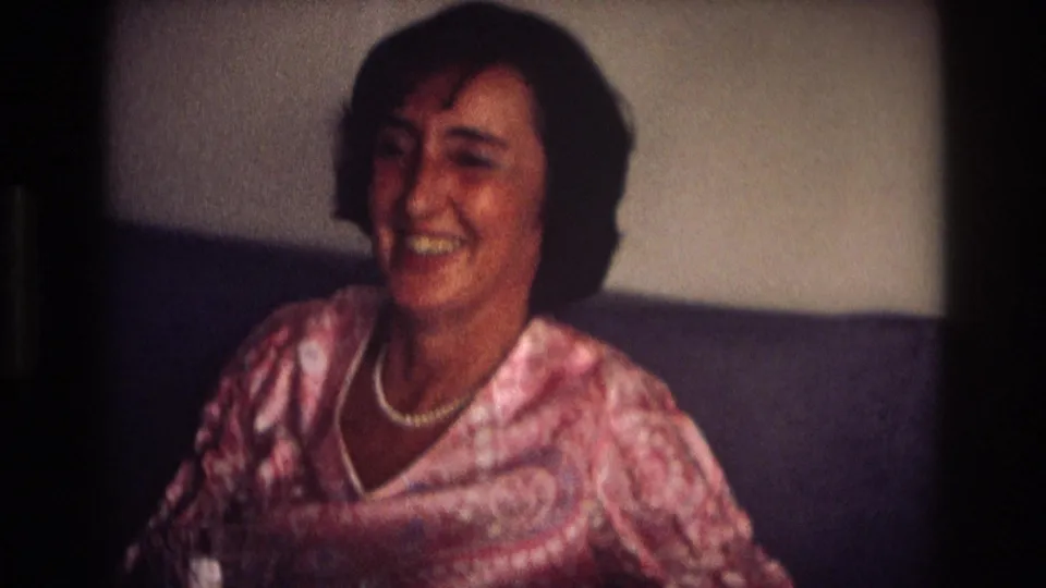 In 1968 Ocean City home-movie footage, a woman wearing a pink patterned blouse and white necklace smiles toward the camera indoors.