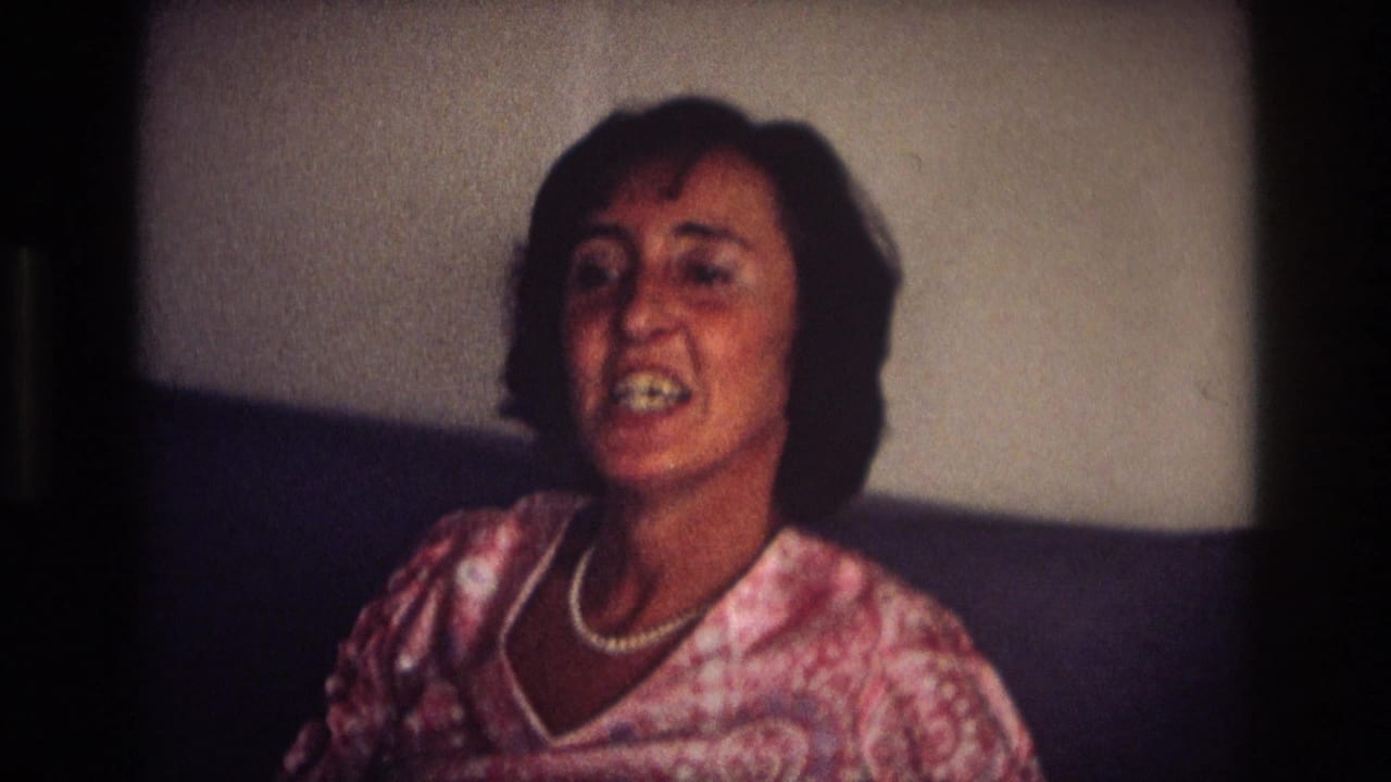 The woman in a pink blouse and white necklace faces the camera, her expression shifting as if speaking in the 1968 indoor home-movie scene.