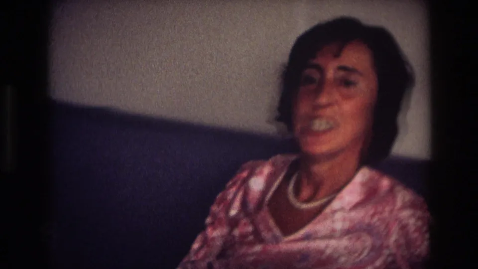A softly focused frame shows the woman seated indoors on a dark couch, wearing a pink patterned blouse and white necklace.