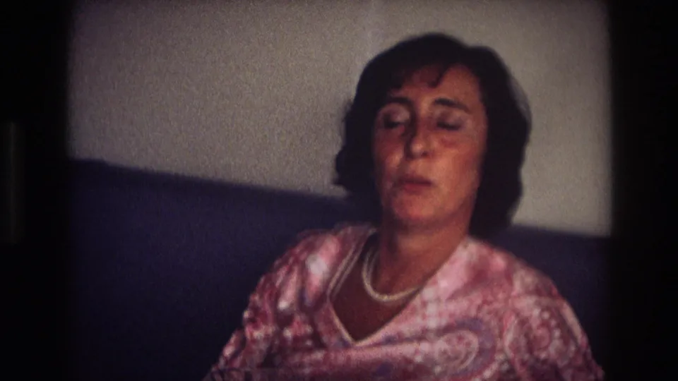 The 1968 home-movie frame shows the woman on a dark couch, looking down or mid-speech while wearing a white necklace and pink blouse.