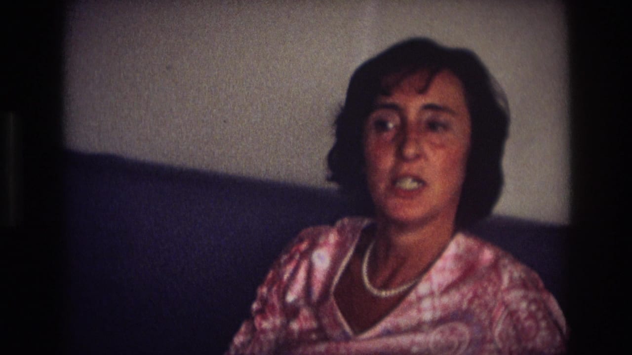 Adult woman in a patterned dress and pearl necklace sits on a dark couch in a 1968 Ocean City, New Jersey home-movie interior.
