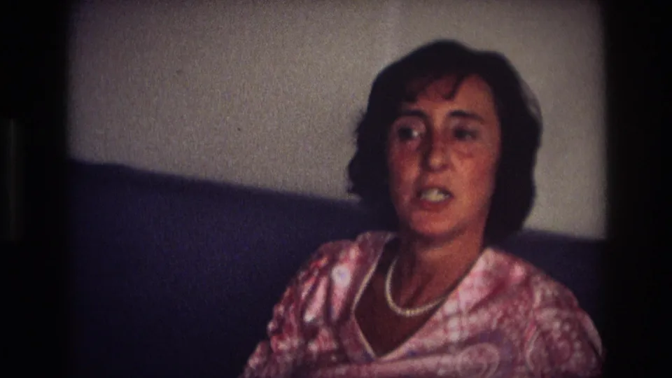 Adult woman in a patterned dress and pearl necklace sits on a dark couch in a 1968 Ocean City, New Jersey home-movie interior.