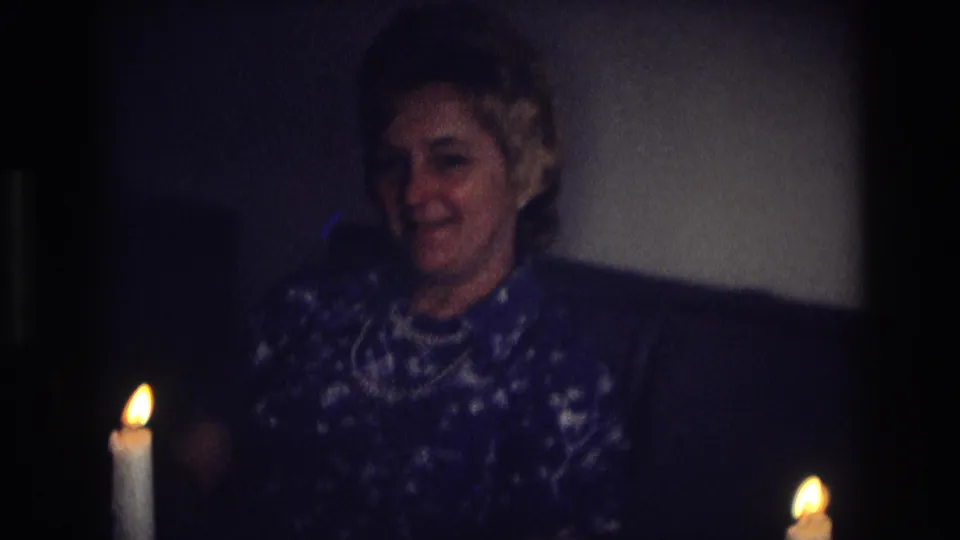Adult woman in a patterned blue dress and pearls sits on a couch beside candlelight in the supplied 1968 Ocean City home-movie setting.