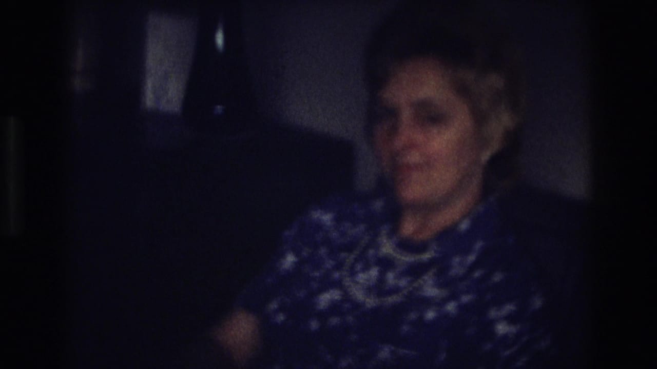 Older woman in 1968 Ocean City home-movie footage smiles in a low-lit private interior, wearing a blue patterned dress and pearl necklace.