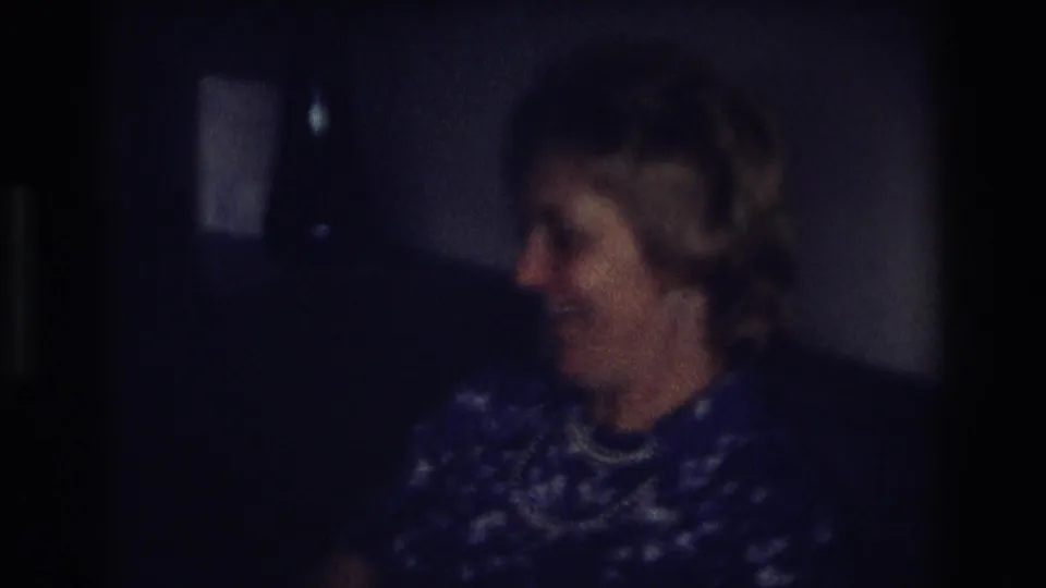 Older woman in the 1968 Ocean City home movie turns sideways and smiles, with her pearl necklace visible against a blue patterned dress.
