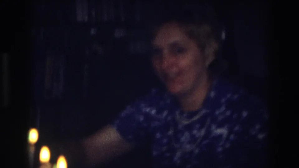 Older woman in 1968 Ocean City home-movie footage leans forward in a grainy, blue-toned indoor frame.