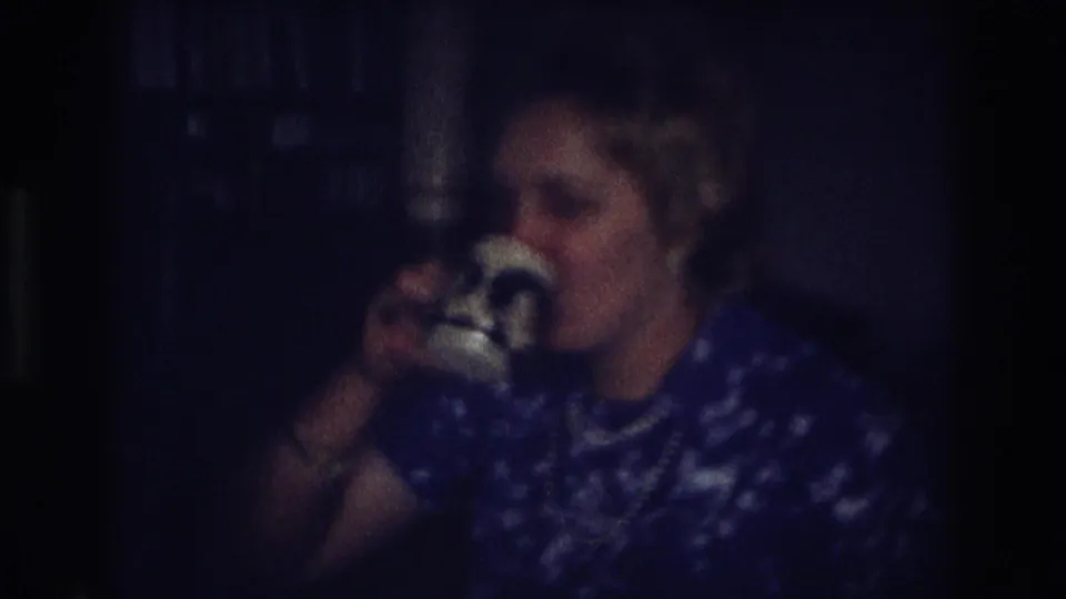 Older woman in 1968 Ocean City home-movie footage lifts a white cup to drink while seated in a dim private interior.