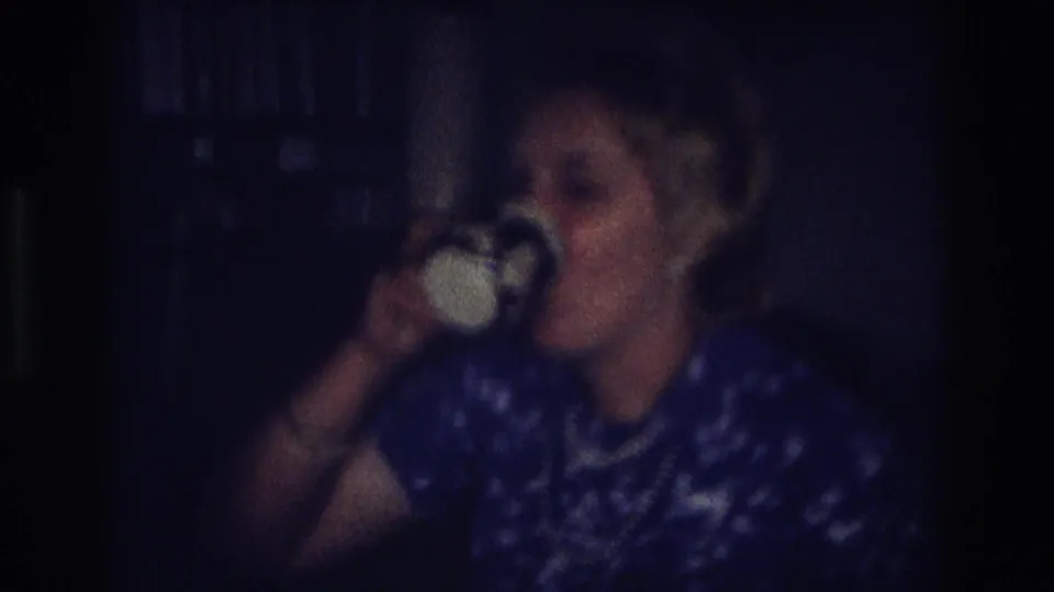 In a 1968 Ocean City, New Jersey home-movie frame, a woman in a blue patterned dress sips from a white cup in a dim interior.