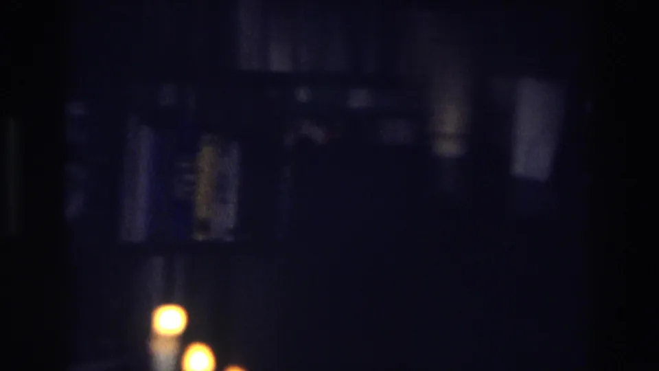 A dim home-movie frame shows a candle flame in the foreground with dark shelves or household objects behind it.