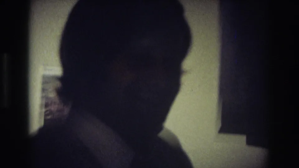 A silhouetted adult man in the 1968 Ocean City interior passes through low light, with a bright wall or doorway behind his head and shoulder.