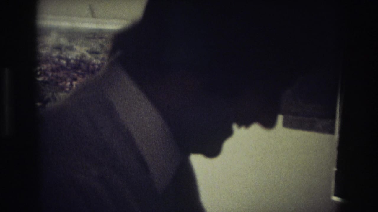In 1968 Ocean City home-movie footage, a man in a dark suit and tie leans in profile inside a shadowed kitchen.