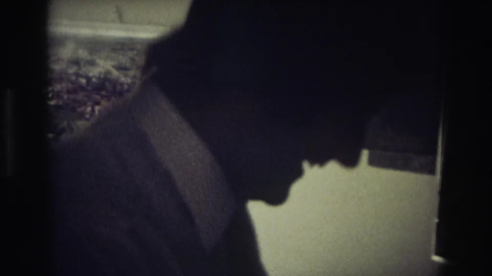 In 1968 Ocean City home-movie footage, a man in a dark suit and tie leans in profile inside a shadowed kitchen.