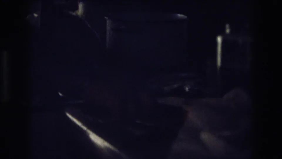 A close, underexposed 8mm frame shows a hand using a knife near bread in a private kitchen interior.