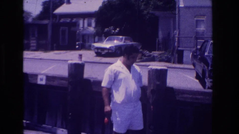 1968 Ocean City boardwalk frame with a man in white summer clothes, wood posts, parked cars, and houses across the street.